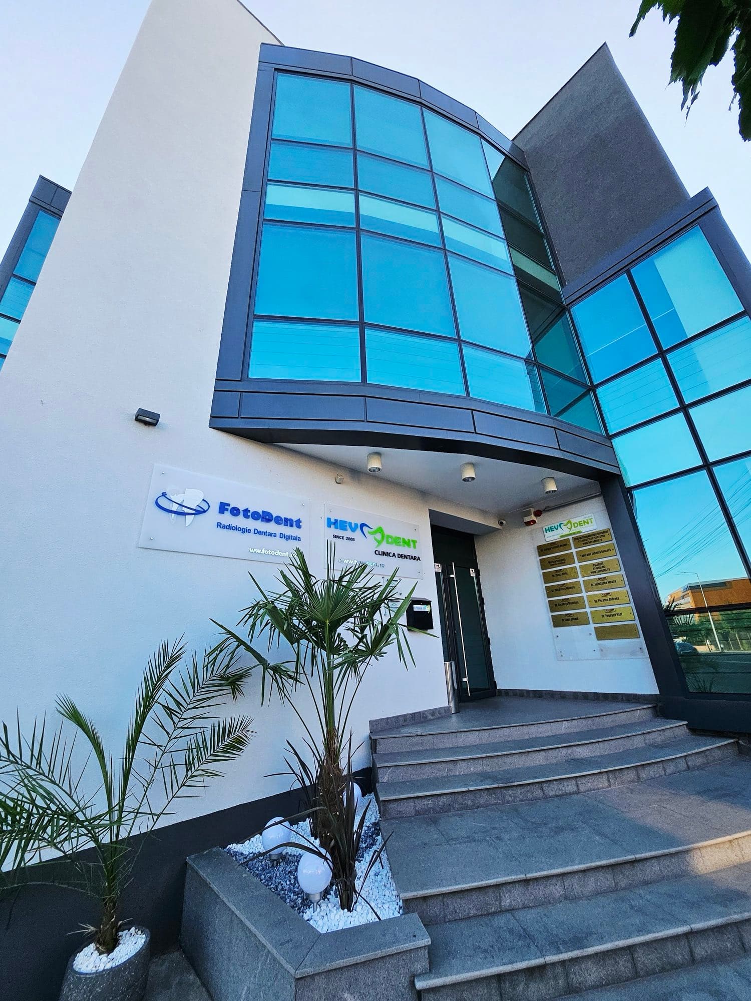 The facade of the Hevadent building in Targu Jiu, with a curved blue glass window, the FotoDent and Hevadent signs and the entrance steps